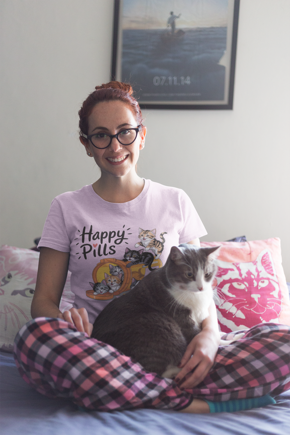 Woman in glasses wearing a lilac Happy Pills t-shirt, sitting in bed with her cat in a cozy bedroom setting