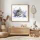 May include: Watercolor painting of a kitten and a mouse in a wooden frame. The kitten has green eyes and a brown and grey coat. The mouse is grey. The background has purple watercolor splashes. The artwork is displayed in a room with a woven cabinet and a teddy bear.