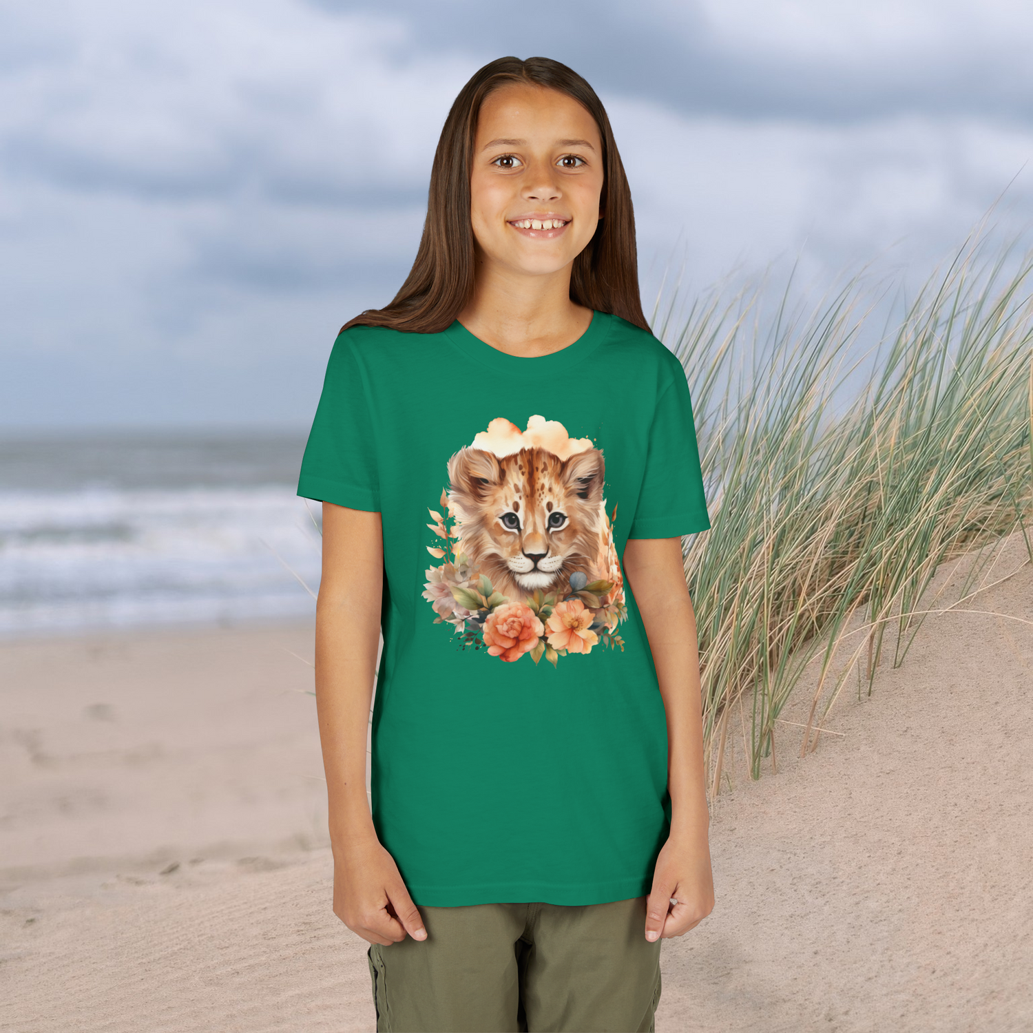 Happy girl in a vibrant kelly green Lion Cub Floral Youth T-Shirt with a watercolor lion cub and colorful flowers, ocean and dunes behind
