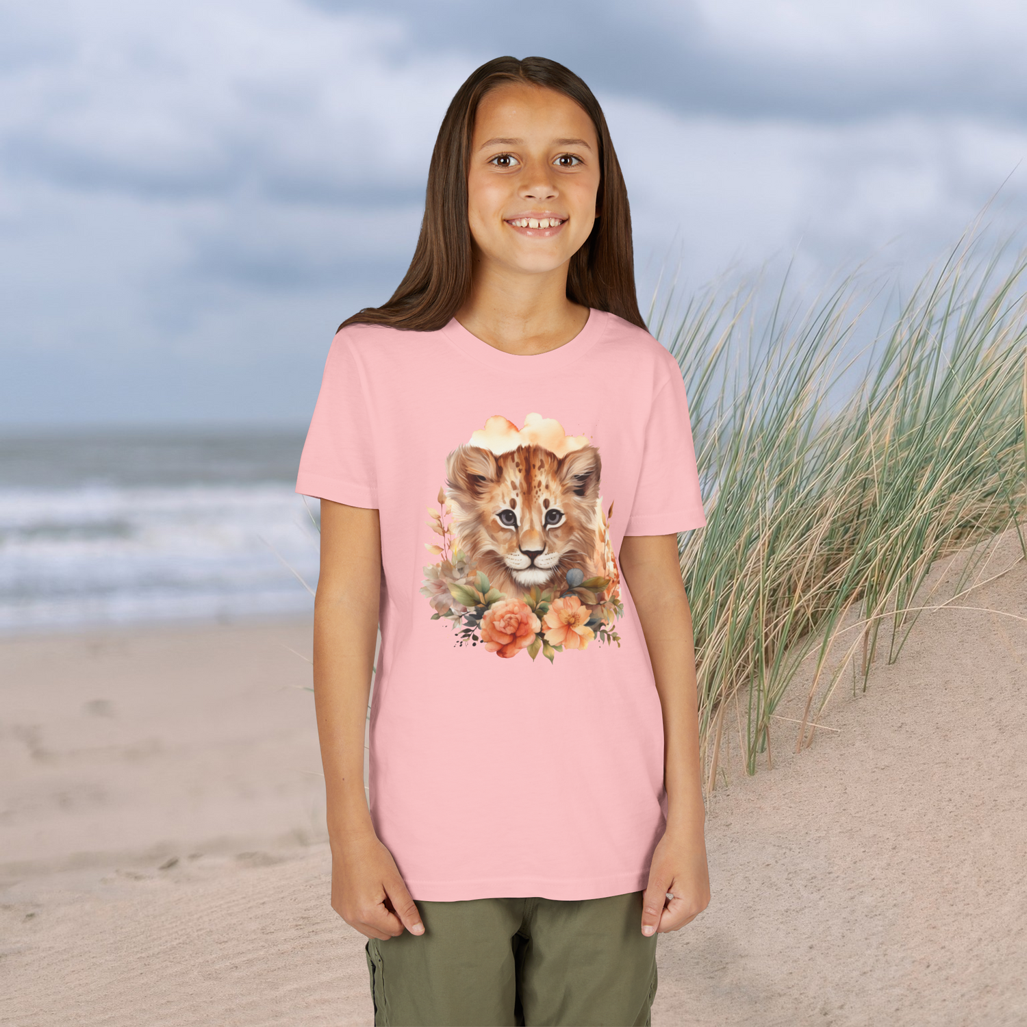 Smiling young girl in a pink Lion Cub Floral Youth T-Shirt with a cute lion cub and floral artwork, beach background
