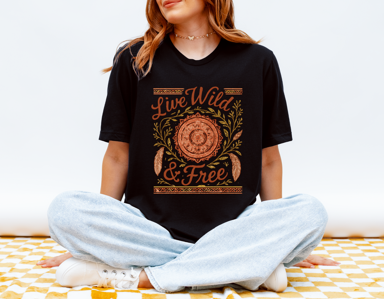 Woman wearing the Live Wild & Free Boho Mandala Feather T-Shirt in black, sitting cross-legged on a yellow checkered blanket — a soft, ethically made free spirit tee from Peaceful Nature Designs