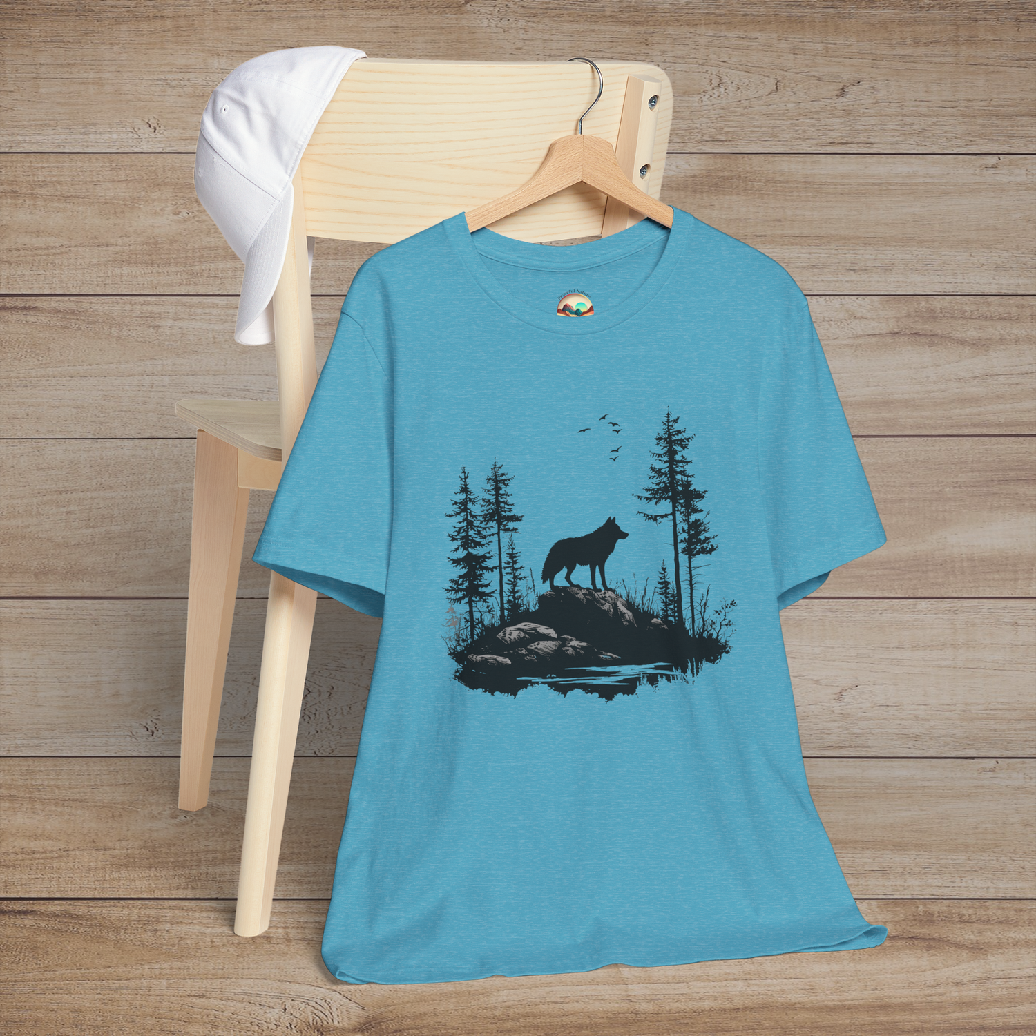 Heather aqua t-shirt displaying a powerful lone wolf standing on a rocky terrain surrounded by pine trees and soaring birds, celebrating resilience