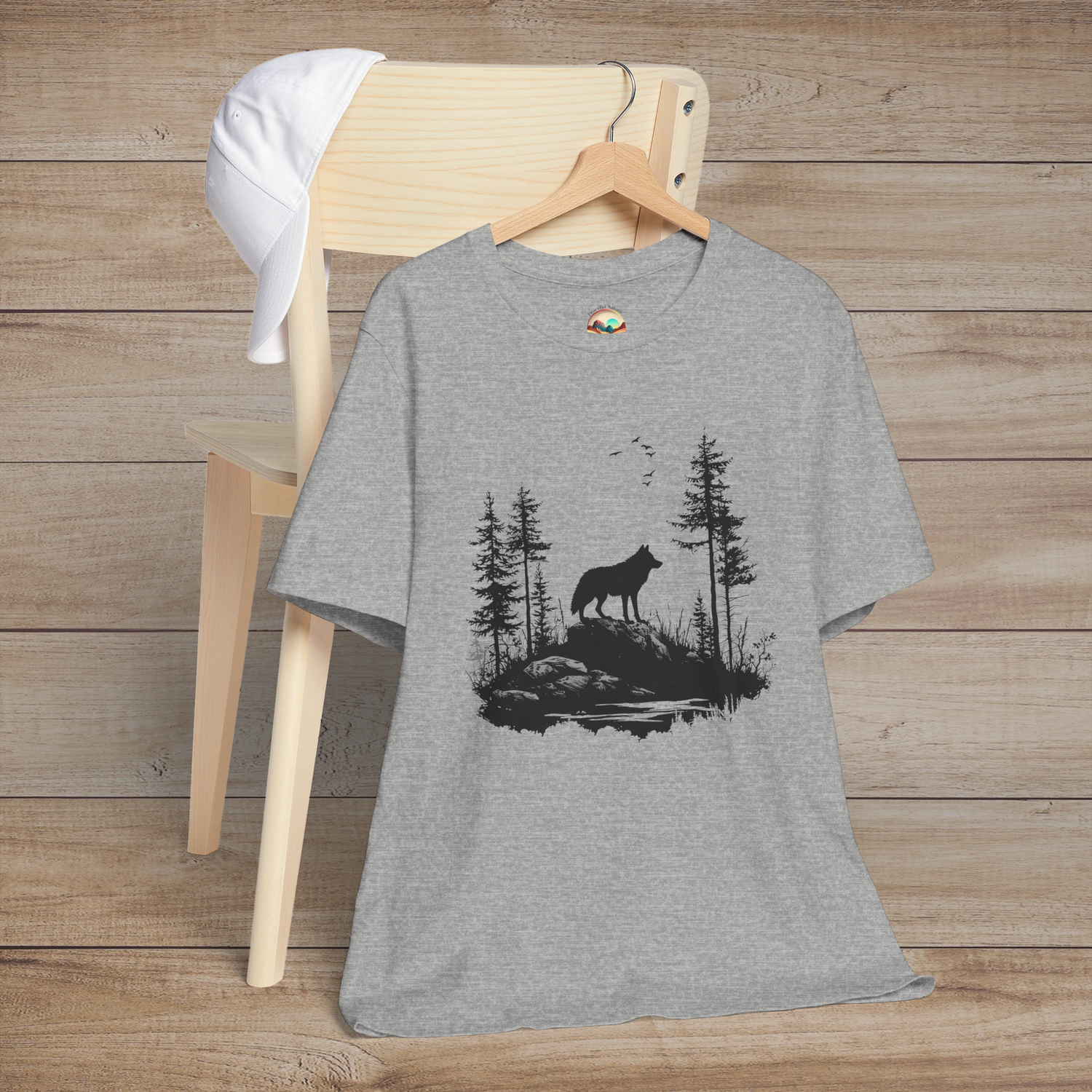 Heather grey t-shirt featuring a lone wolf howling on a rocky cliff surrounded by evergreen trees and flying birds, symbolizing strength and independence