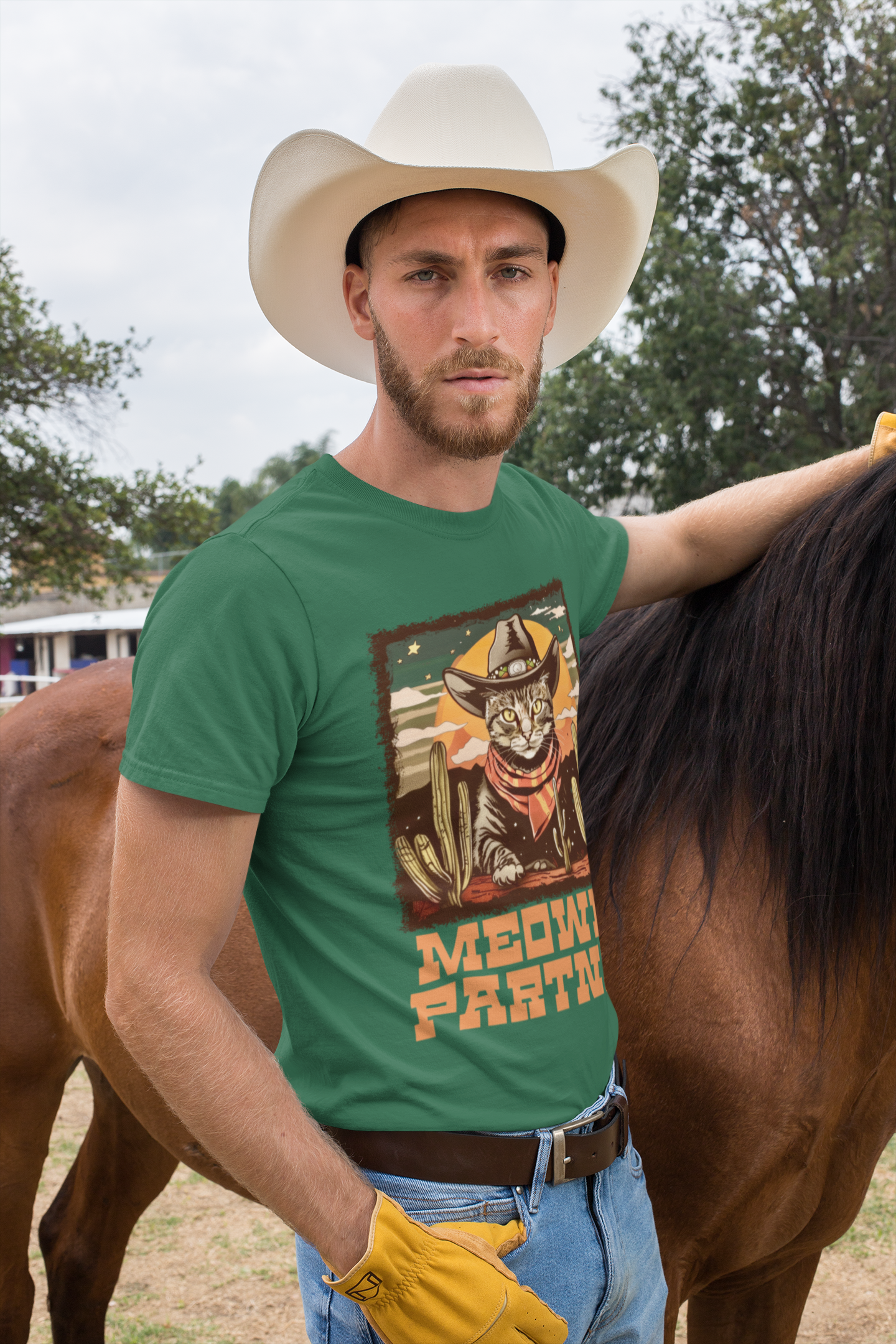 Male model with cowboy hat standing beside a horse wearing the Heather Green Grass Meowdy Partner Cowboy Cat Tee, showing a cat with Wild West style