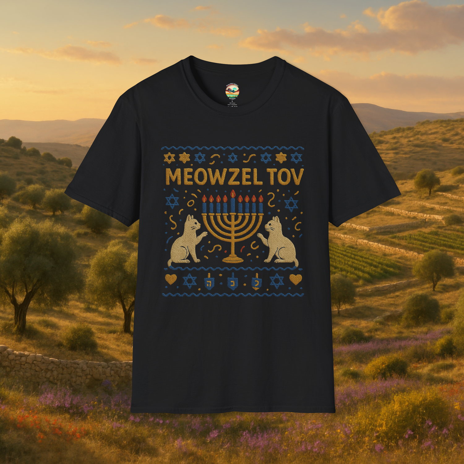 Flat lay of a black t-shirt featuring a whimsical embroidered-style "Meowzel Tov" Hanukkah design with two cat silhouettes, a nine-candle menorah, Stars of David, dreidels, and confetti, set against a scenic Mediterranean landscape background