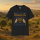 Flat lay of a black t-shirt featuring a whimsical embroidered-style "Meowzel Tov" Hanukkah design with two cat silhouettes, a nine-candle menorah, Stars of David, dreidels, and confetti, set against a scenic Mediterranean landscape background