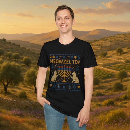 Smiling man wearing a black "Meowzel Tov" Hanukkah cat t-shirt with an embroidered-style menorah, cats, and festive symbols, standing in front of a scenic Mediterranean landscape at sunset