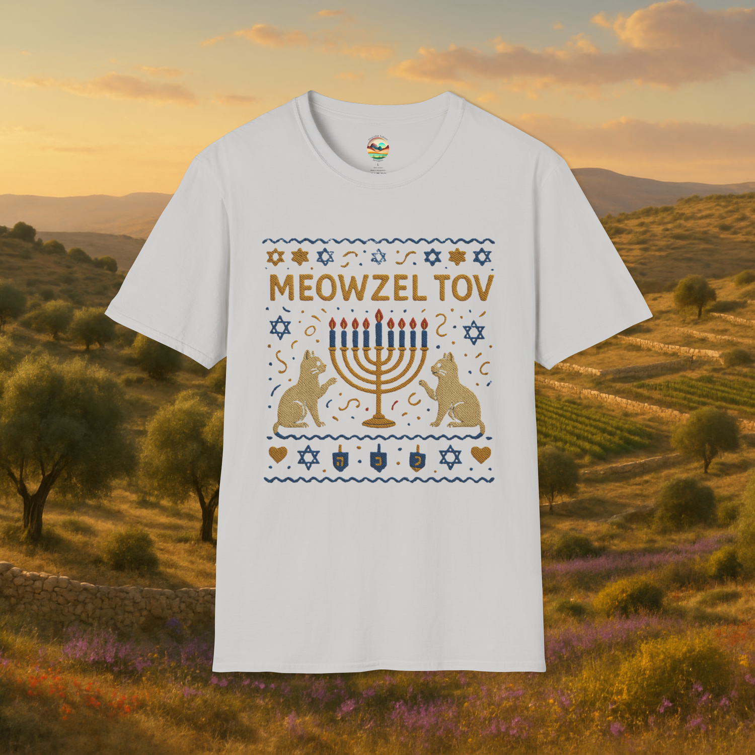 Flat lay of an ice grey t-shirt displaying a festive "Meowzel Tov" Hanukkah cat design with cats, menorah, dreidels, and Stars of David, placed against a rustic Mediterranean field backdrop