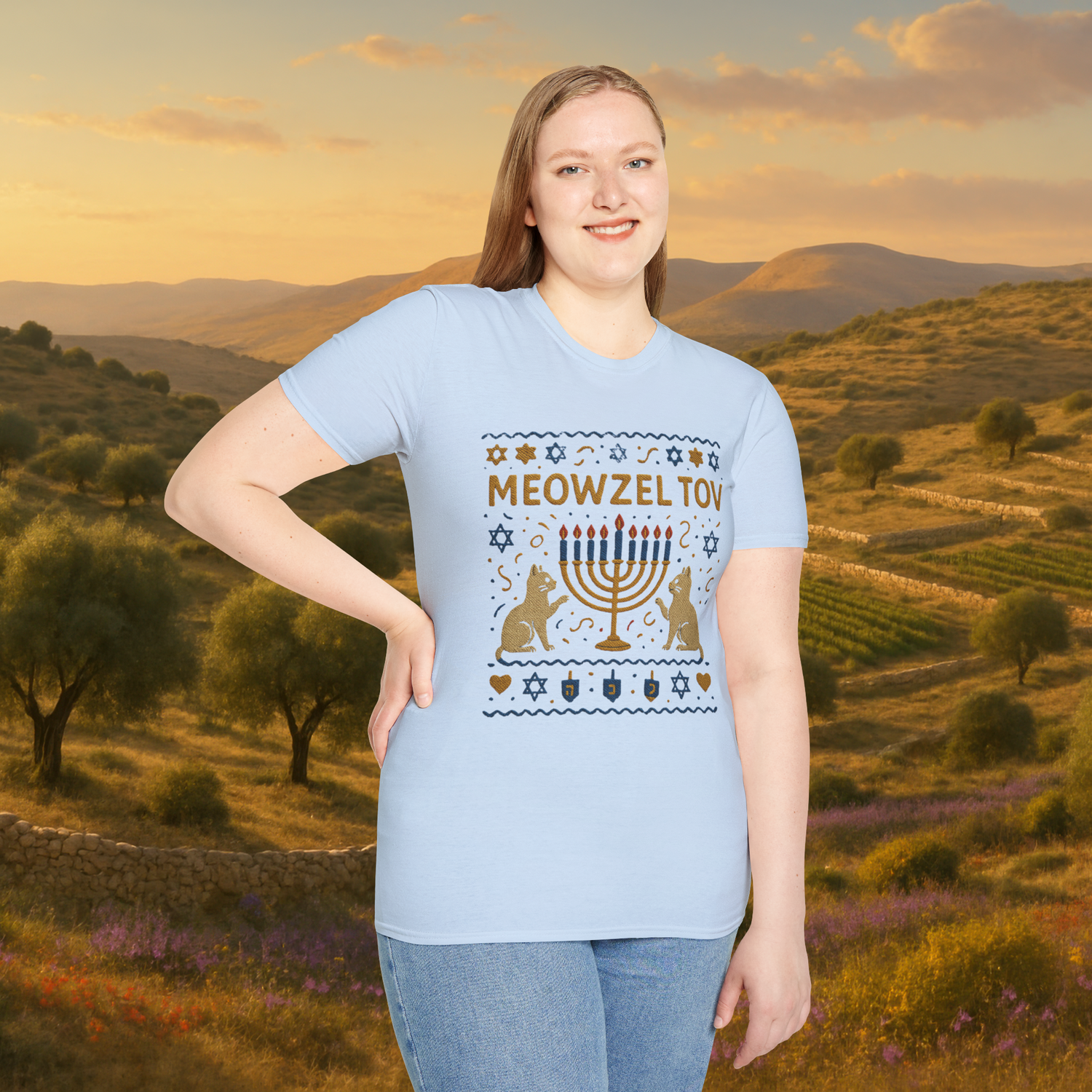 Woman posing in a light blue "Meowzel Tov" Hanukkah cat t-shirt, featuring an embroidered-style menorah and festive cat-themed holiday graphics, with a backdrop of rolling hills and olive trees