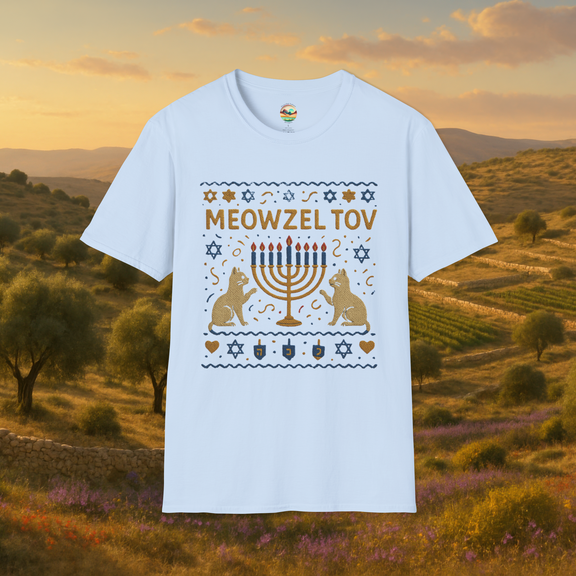 Flat lay of a light blue t-shirt showing a "Meowzel Tov" Hanukkah cat design with embroidered-style cats, a nine-candle menorah, dreidels, and Stars of David, set against a natural field landscape at sunset