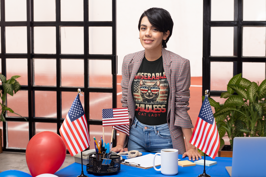 Woman standing behind desk with American flags and balloons, wearing the "Miserable Childless Cat Lady" soft fit black t-shirt