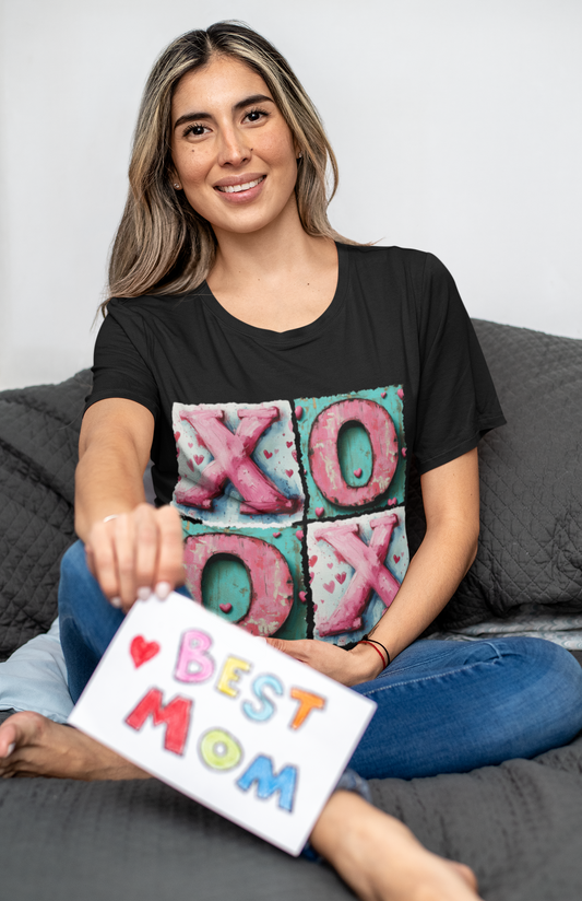 Smiling woman sitting on a couch wearing a black Hugs and Kisses T-Shirt, holding a handmade “Best Mom” card, perfect for Mother’s Day or Valentine’s Day