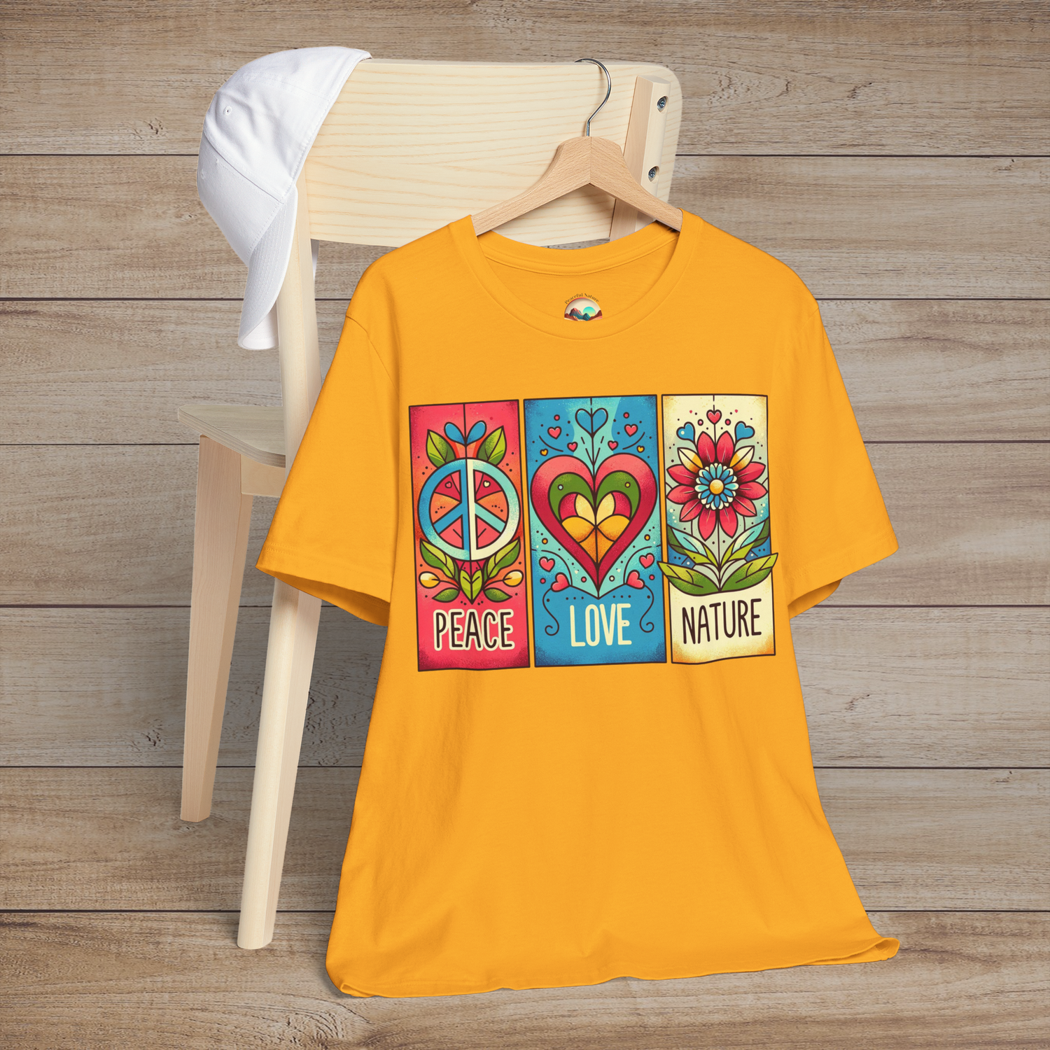 Gold variant of the Peace Love and Nature T-Shirt laid on a chair with white hat, featuring a bold design of peace sign, heart, and flower blocks in vivid reds, blues, and greens. A statement piece for outdoor and festival wear
