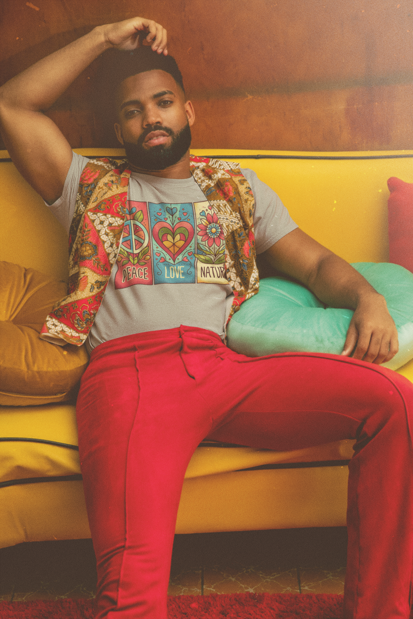 Male model reclining on a yellow couch in heather grey (Athletic Heather) Peace Love Nature t-shirt, paired with red pants and a colorful vest in a retro room