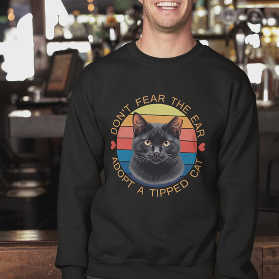 A man wearing a Gildan 18000 sweatshirt with a retro “Don’t Fear the Ear – Adopt a Tipped Cat” black cat design pets a cute cat indoors, showcasing the shirt’s fit and animal-rescue message.
