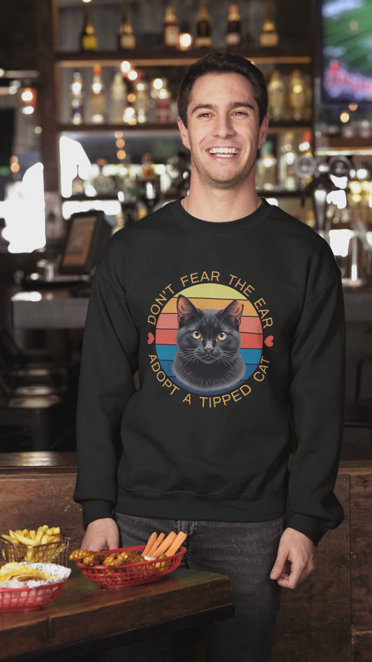 A man wearing a Gildan 18000 sweatshirt with a retro “Don’t Fear the Ear – Adopt a Tipped Cat” black cat design pets a cute cat indoors, showcasing the shirt’s fit and animal-rescue message.