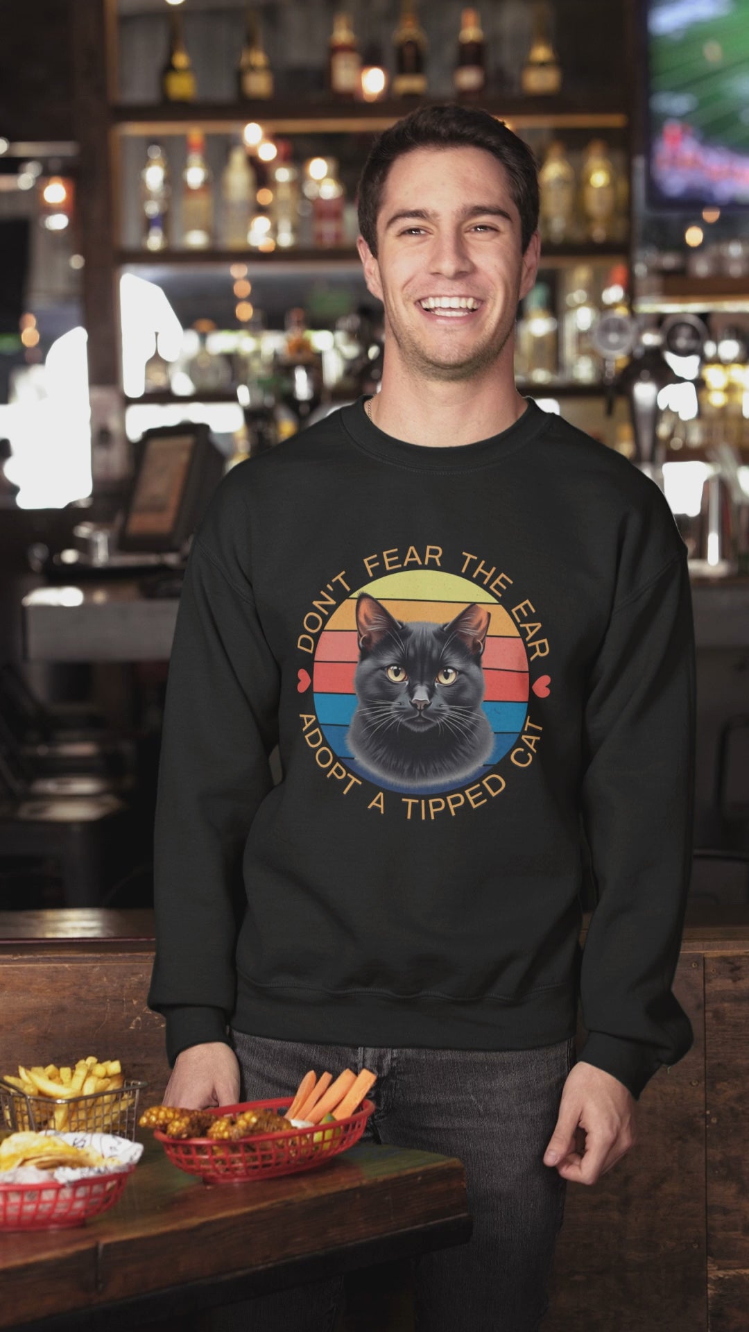 A man wearing a Gildan 18000 sweatshirt with a retro “Don’t Fear the Ear – Adopt a Tipped Cat” black cat design pets a cute cat indoors, showcasing the shirt’s fit and animal-rescue message.