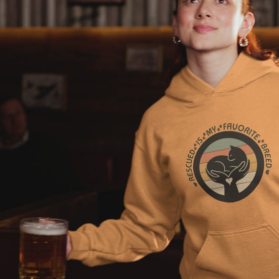 Video of a smiling woman celebrating with a drink while wearing a tangerine ‘Rescued Is My Favorite Breed’ retro cat rescue hoodie