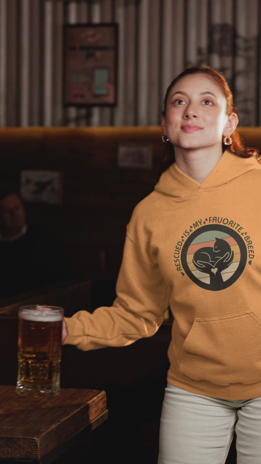 Video of a smiling woman celebrating with a drink while wearing a tangerine ‘Rescued Is My Favorite Breed’ retro cat rescue hoodie