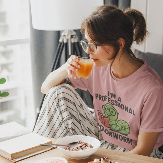 Video of woman wearing the heather mauve t-shirt with a graphic and text 'I'm a Professional Bookworm' on a wooden chair.