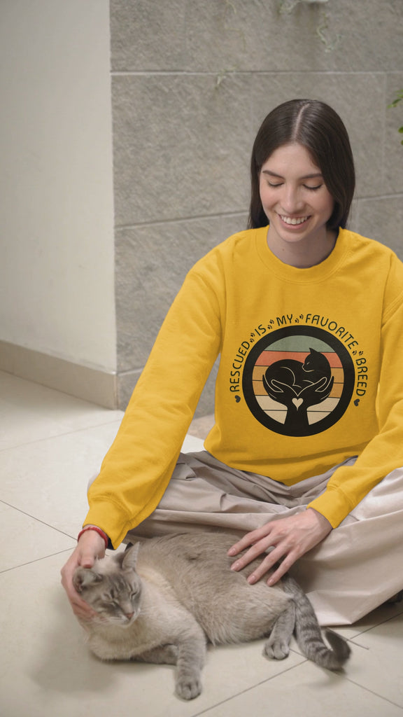 Video of woman who is wearing the Rescued is My Favorite Breed Gildan 18000 sweatshirt in Gold. The woman is petting a cat. 
