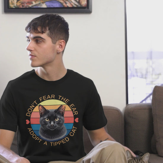 A man wearing a Bella Canvas t-shirt with a retro “Don’t Fear the Ear – Adopt a Tipped Cat” black cat design pets a cute cat indoors, showcasing the shirt’s fit and animal-rescue message