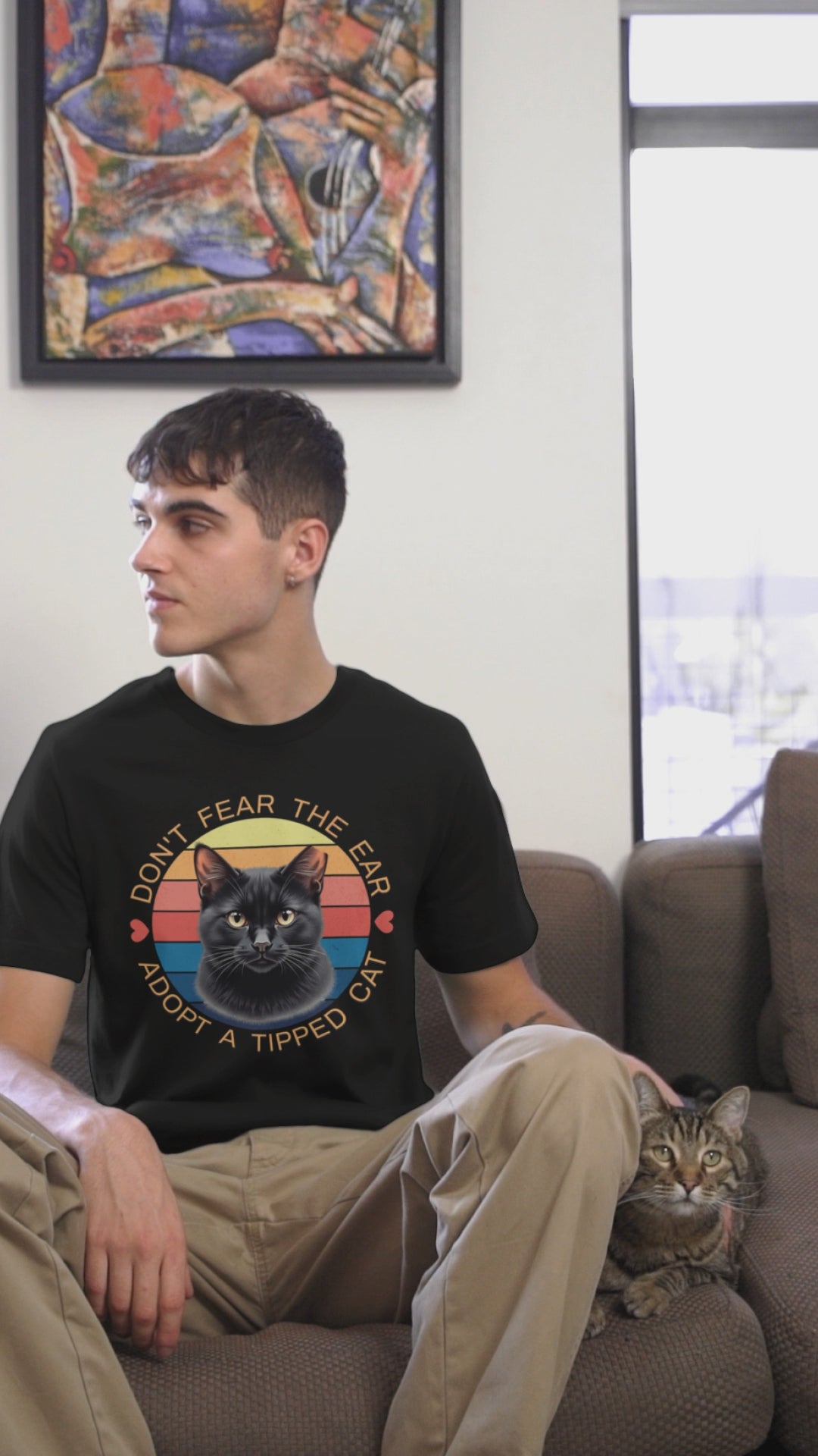 A man wearing a Bella Canvas t-shirt with a retro “Don’t Fear the Ear – Adopt a Tipped Cat” black cat design pets a cute cat indoors, showcasing the shirt’s fit and animal-rescue message