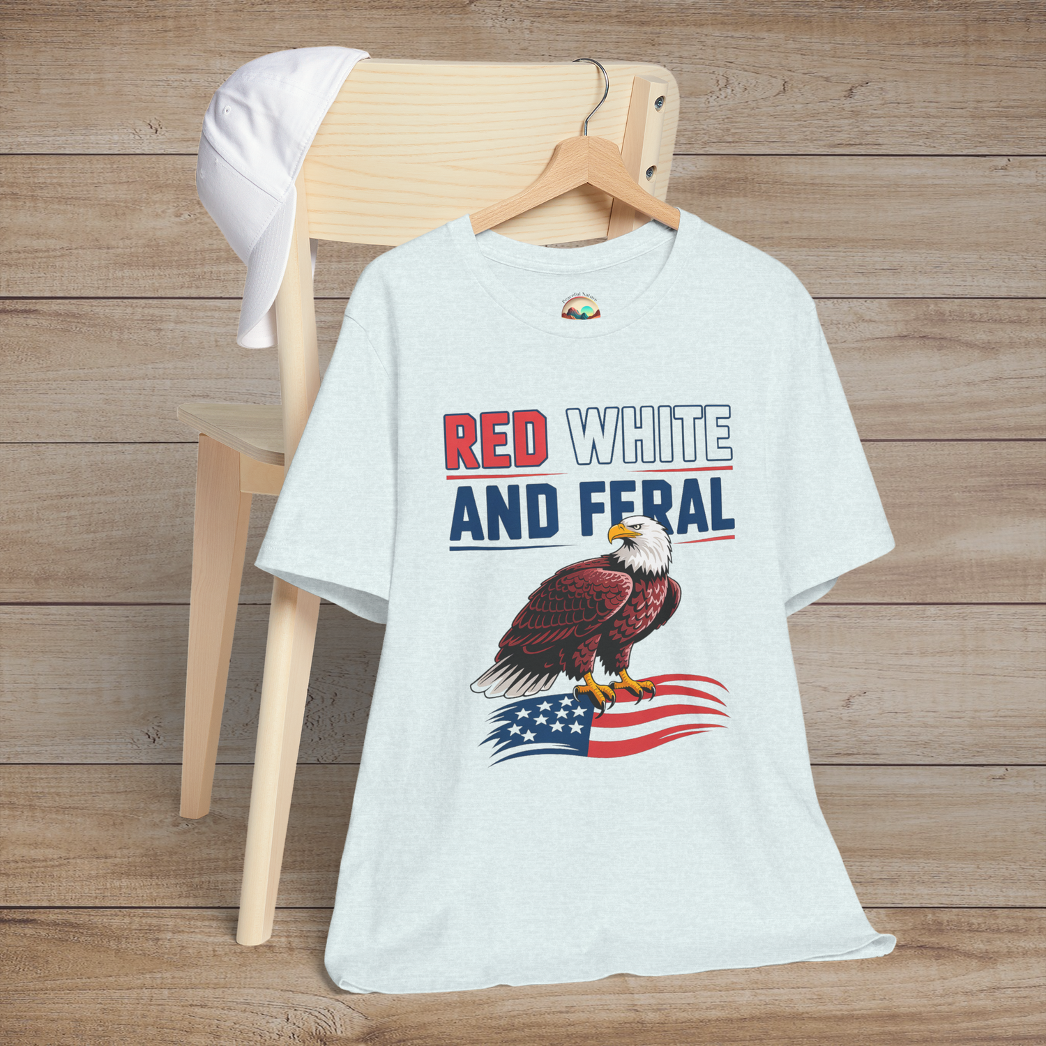 Fourth of July-themed graphic tee in heather ice blue with a fierce bald eagle and the text "Red White and Feral" in bold patriotic colors.