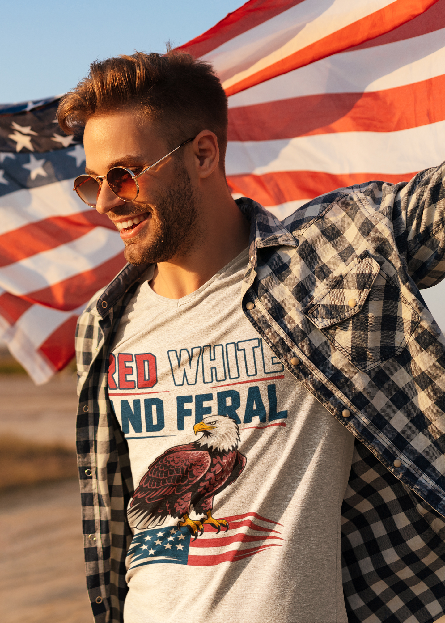 Smiling man wearing a "Red White and Feral" eagle t-shirt while holding an American flag outdoors, celebrating patriotic spirit with casual style.