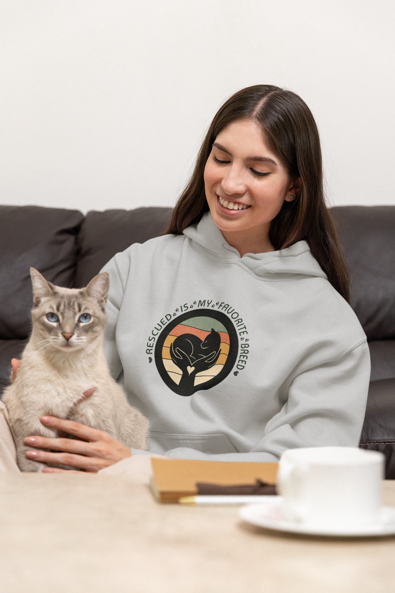 Smiling woman wearing a light gray 'Rescued Is My Favorite Breed' retro cat rescue hoodie while holding her cat on the couch