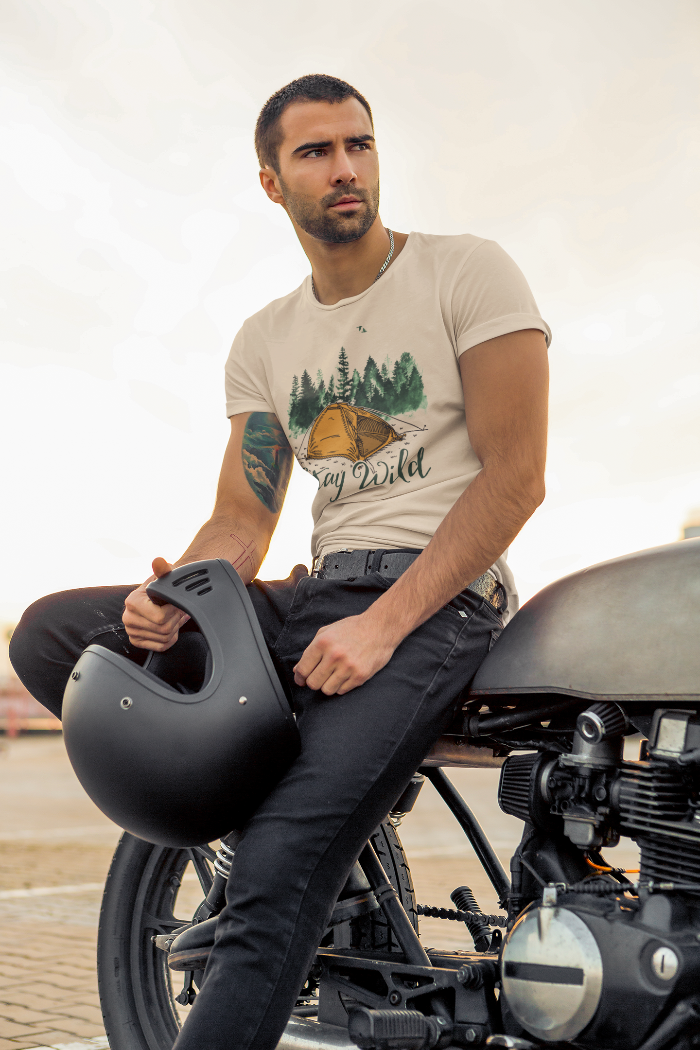 A rugged man with short dark hair and a trimmed beard sits confidently on a motorcycle, wearing a light-colored "Stay Wild" t-shirt featuring an orange tent and pine trees. He holds a matte black helmet in one hand and looks into the distance with a thoughtful expression, embodying a bold and adventurous spirit.
