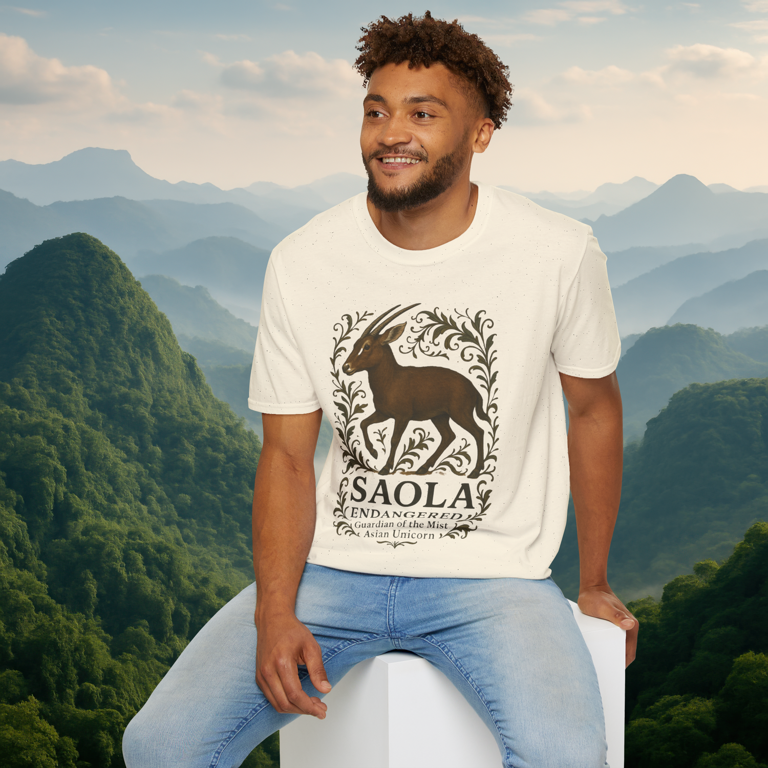 Man seated and wearing the Saola Endangered Asian Unicorn T-Shirt in Natural – eco-friendly cotton tee featuring Saola art and foliage, with misty forested mountains behind him