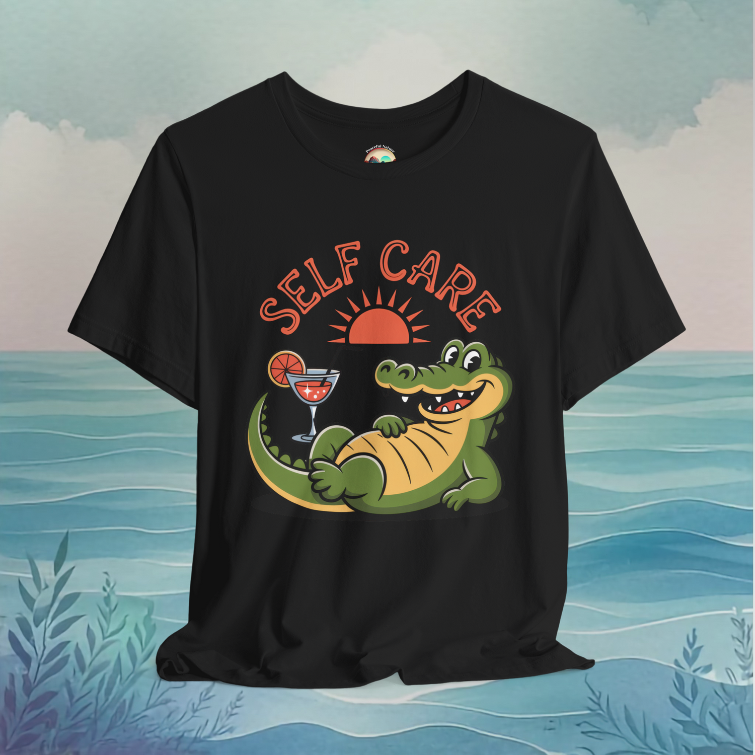 Black t-shirt with relaxed cartoon alligator, red sun, drink glass, and playful "SELF CARE" typography