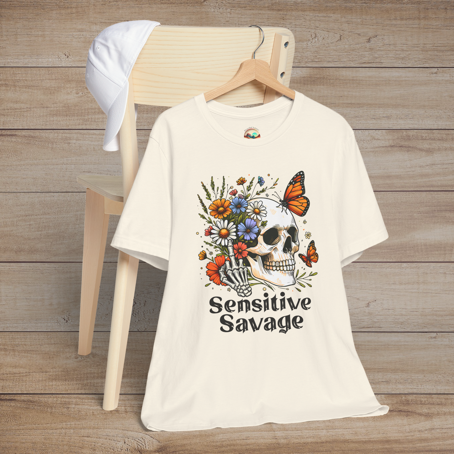 Natural-colored t-shirt featuring bold Sensitive Savage text with a skull holding blooming flowers and surrounded by butterflies