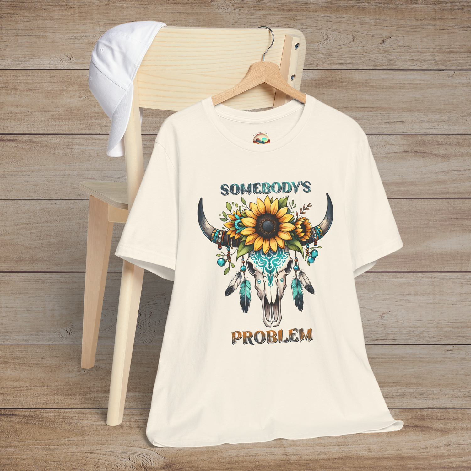 Somebody’s Problem T-Shirt with a vibrant Boho skull design adorned with flowers and turquoise feathers on natural color fabric