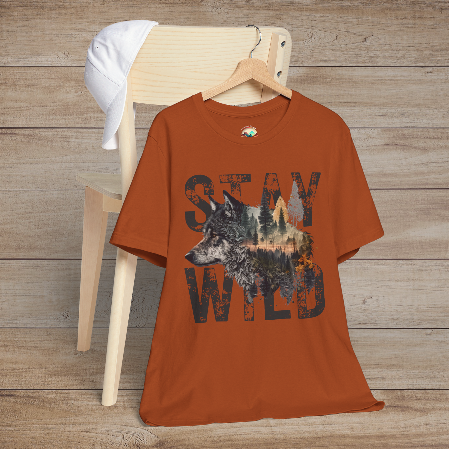 Burnt orange "Stay Wild" T-shirt with bold black lettering and a majestic wolf illustration blended into a forest landscape, displayed flat on a wooden chair with a white baseball cap