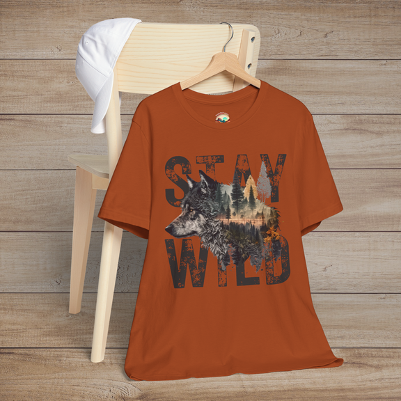Burnt orange "Stay Wild" T-shirt with bold black lettering and a majestic wolf illustration blended into a forest landscape, displayed flat on a wooden chair with a white baseball cap