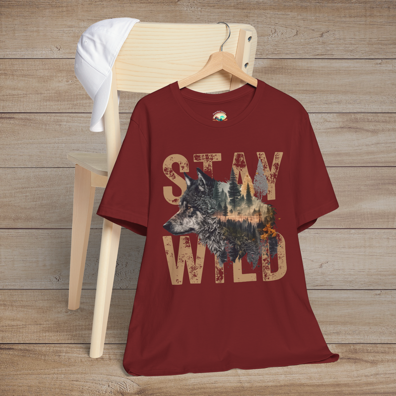 Deep red "Stay Wild" T-shirt featuring a rustic wolf and forest silhouette inside distressed tan letters, styled on a wooden chair for a rugged, nature-inspired look