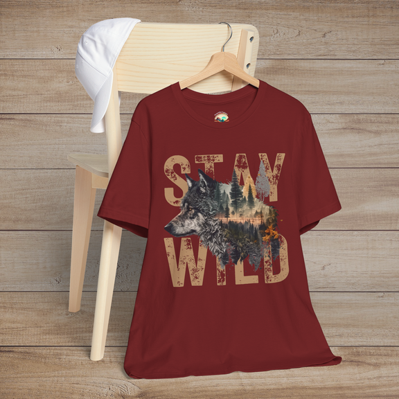Deep red "Stay Wild" T-shirt featuring a rustic wolf and forest silhouette inside distressed tan letters, styled on a wooden chair for a rugged, nature-inspired look