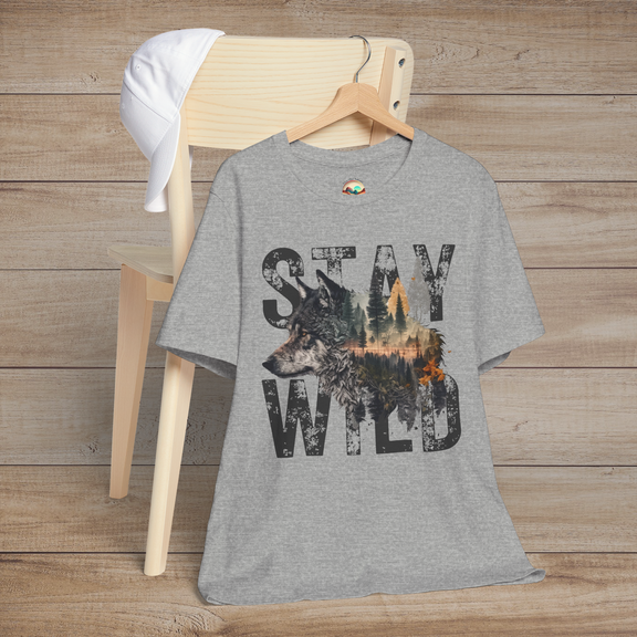 Heather grey (athletic heather) "Stay Wild" T-shirt with a moody grayscale forest and wolf design within weathered block letters, presented as a flat lay on a wooden background