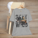 Heather grey (athletic heather) "Stay Wild" T-shirt with a moody grayscale forest and wolf design within weathered block letters, presented as a flat lay on a wooden background