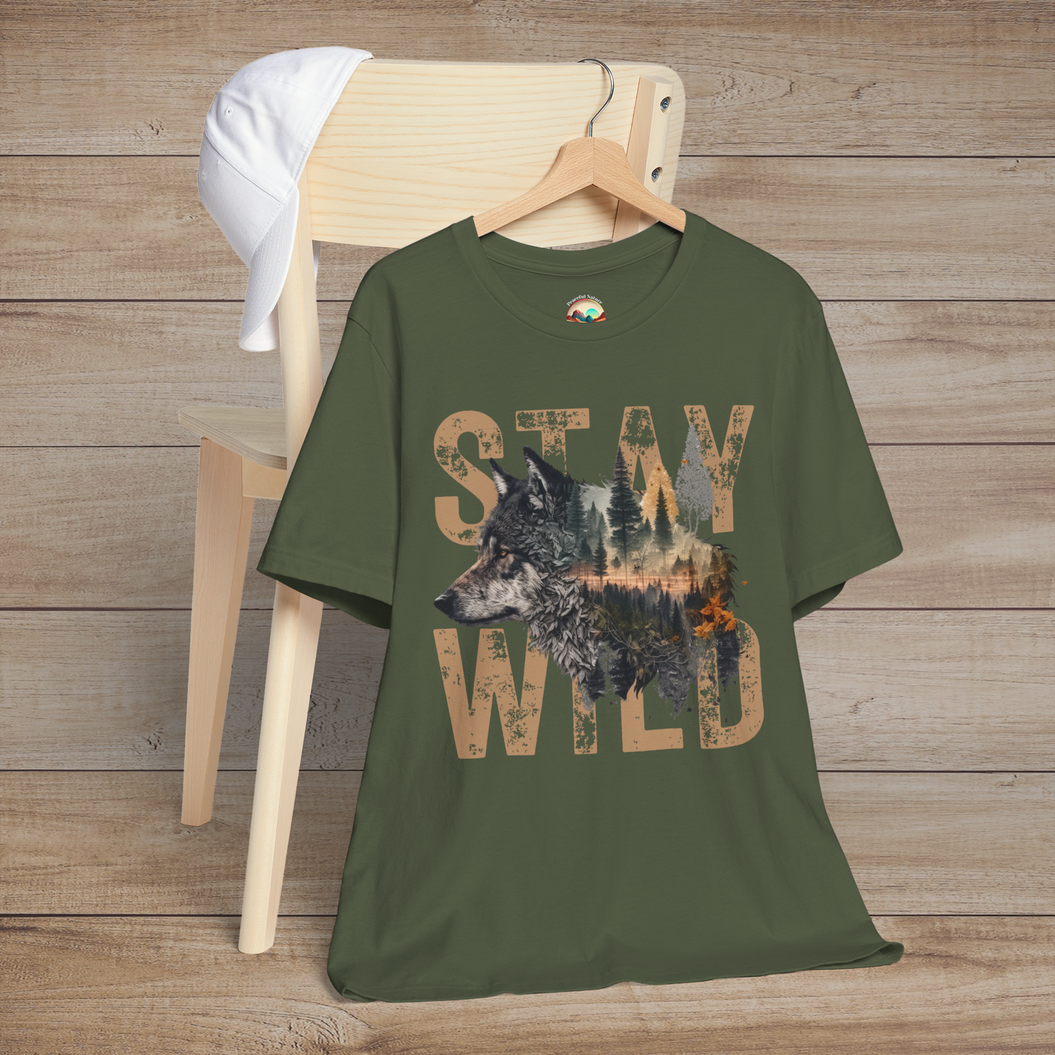 Olive-toned "Stay Wild" T-shirt with a detailed wolf and wilderness scene inside vintage-style tan lettering, laid on a chair with a rustic wooden backdrop