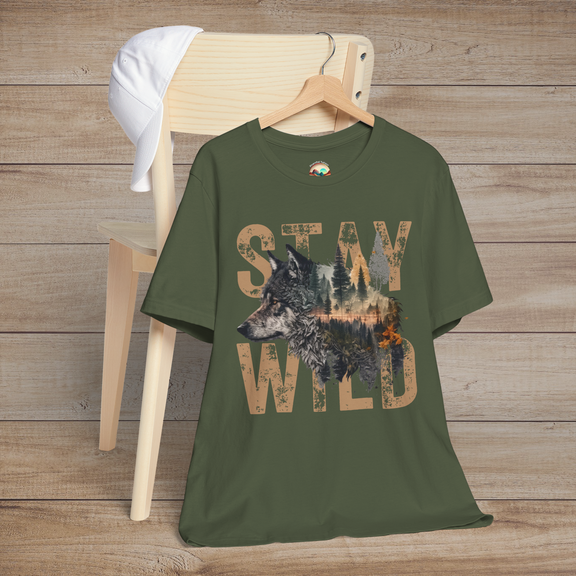 Olive-toned "Stay Wild" T-shirt with a detailed wolf and wilderness scene inside vintage-style tan lettering, laid on a chair with a rustic wooden backdrop