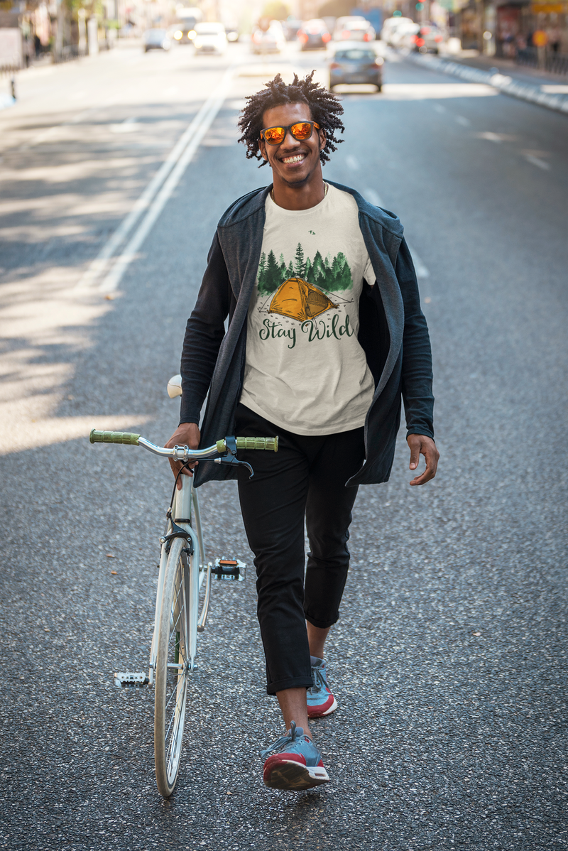 A smiling man in sunglasses walks his bicycle down a sunlit city street, wearing a light "Stay Wild" t-shirt with an illustration of a camping tent and forest. He’s dressed in a casual hoodie and sneakers, radiating laid-back energy and urban adventure vibes.