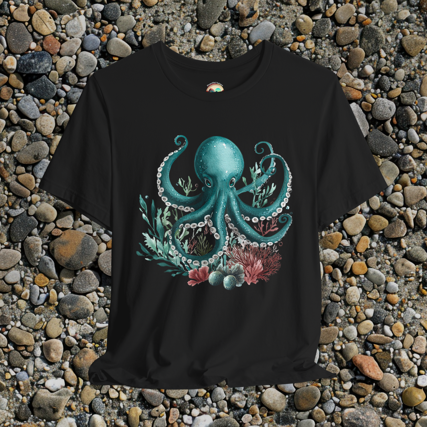 Teal watercolor octopus with coral and seaweed design on a black unisex t-shirt, displayed on a bed of pebbles