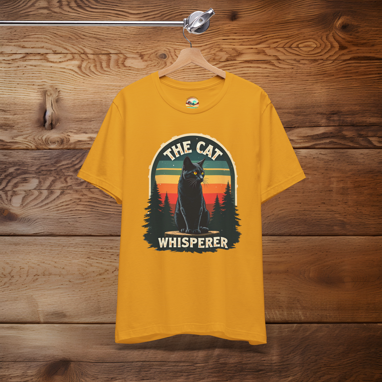 The Cat Whisperer T-Shirt featuring a mystical black cat sitting on a log, surrounded by pine trees and a retro sunset, displayed on a mustard yellow shirt against a rustic wood background