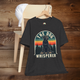 Dark grey heather T-shirt with a vintage retro design featuring a black Labrador sitting on a wooden platform against a colorful sunset and pine tree background. Text reads “The Dog Whisperer.” Shirt is displayed on a wooden hanger draped over a light wood chair with a white cap resting on top.