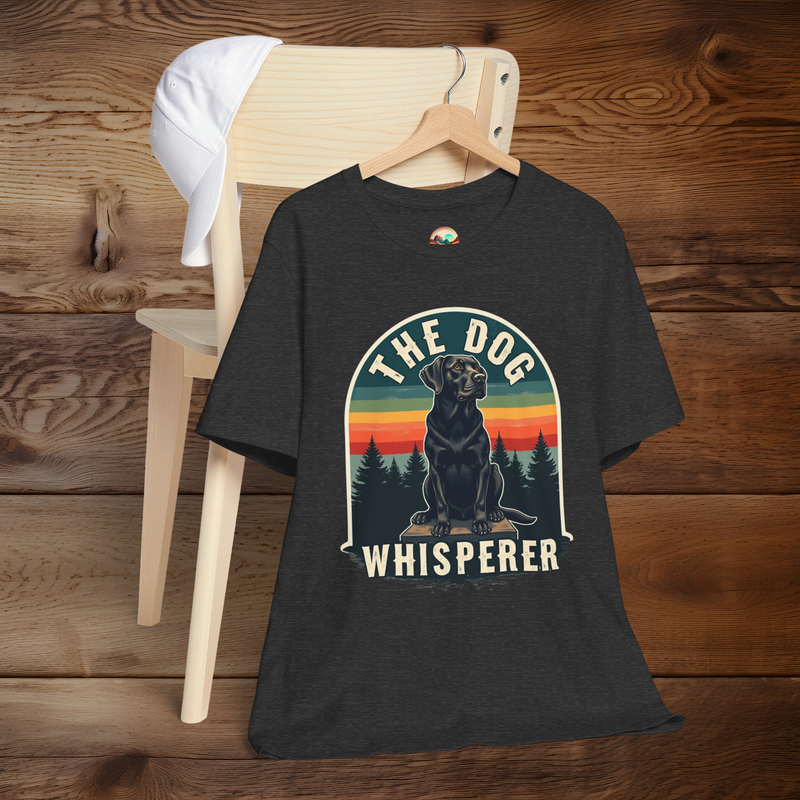 Dark grey heather T-shirt with a vintage retro design featuring a black Labrador sitting on a wooden platform against a colorful sunset and pine tree background. Text reads “The Dog Whisperer.” Shirt is displayed on a wooden hanger draped over a light wood chair with a white cap resting on top.