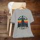 Heather grey T-shirt with a retro-style graphic of a black Labrador sitting in front of a pine tree silhouette and striped sunset. Bold text above and below the dog reads “The Dog Whisperer.” Displayed on a hanger over a light wood chair with a white cap placed on top.
