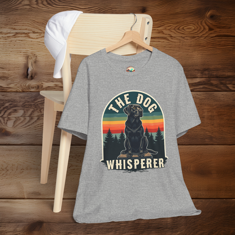 Heather grey T-shirt with a retro-style graphic of a black Labrador sitting in front of a pine tree silhouette and striped sunset. Bold text above and below the dog reads “The Dog Whisperer.” Displayed on a hanger over a light wood chair with a white cap placed on top.